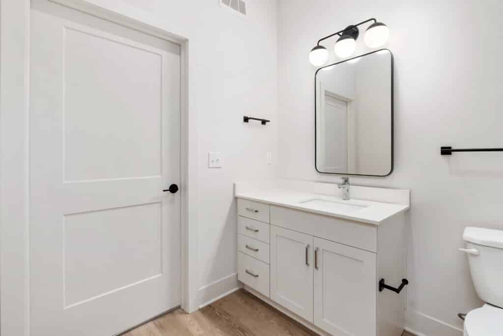 Modern bathroom in a One Bedroom Apartment with a white vanity, rectangular mirror with black frame, three-bulb light fixture, and white walls. Door and toilet visible, featuring sleek black hardware and fixtures.