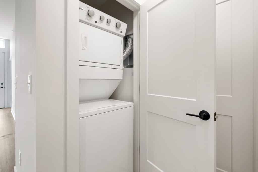 A compact laundry closet in a one bedroom apartment features a white stacked washer and dryer unit, partially enclosed by an open white door.