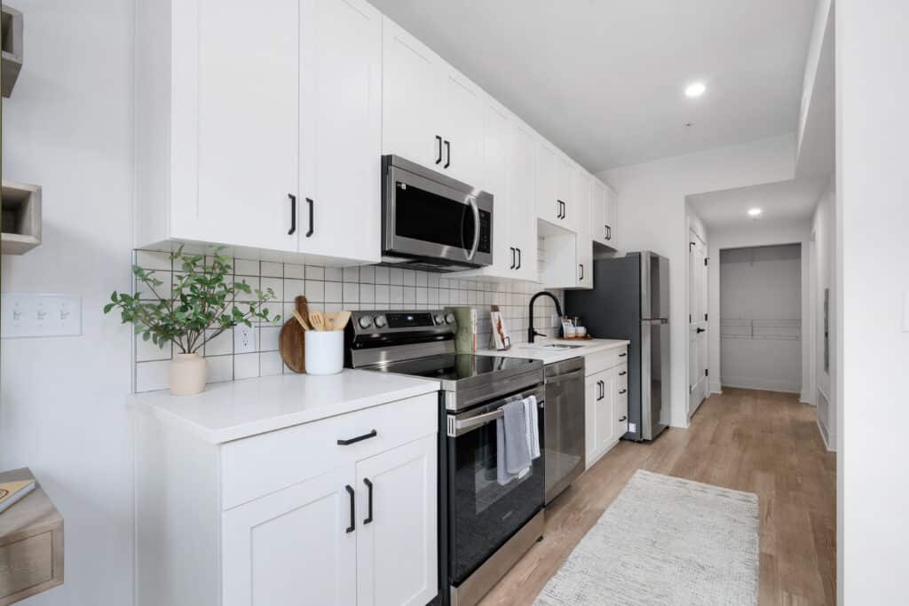 Modern kitchen in a studio apartment with white cabinets, stainless steel appliances, a black faucet, potted plant, and wooden utensils on a light wood floor with a white rug.