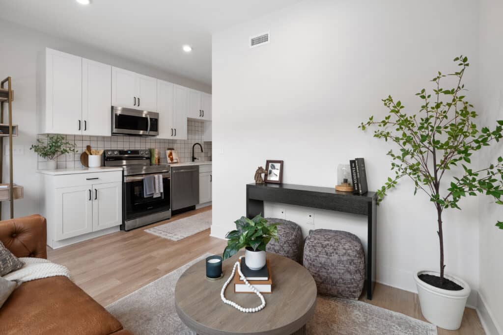 Modern studio apartment interior with a white kitchen, stainless steel appliances, a cozy living area featuring a round coffee table, decorative plants, and minimalist furnishings.