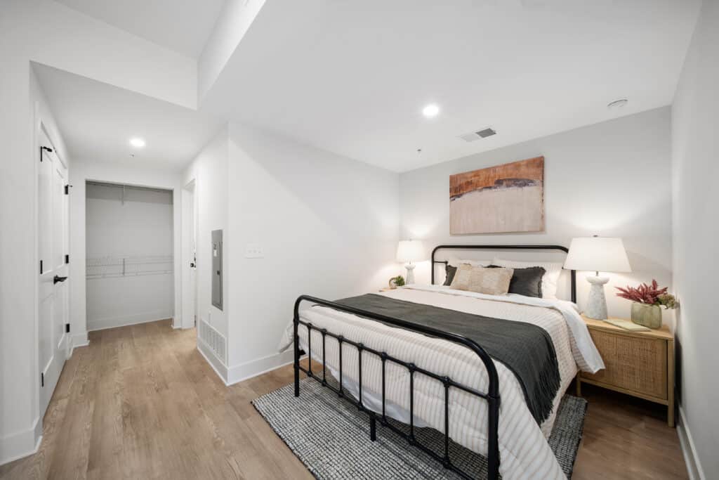 A modern studio apartment bedroom with a black metal bed frame, neutral bedding, two bedside tables with lamps, and a walk-in closet visible in the background.