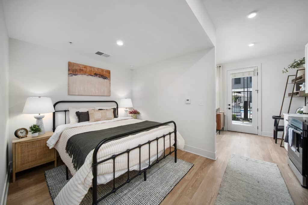 A modern studio apartment bedroom with a black metal bed frame, neutral bedding, two nightstands with lamps, wall art, and a door leading outside. Light wood flooring and white walls complete the airy ambiance.