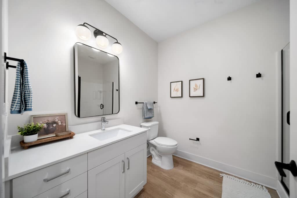 Modern studio apartment bathroom with white walls, wood floor, a single-sink vanity, toilet, wall art, a large mirror, and a glass shower in the background. Towels are hung on black hooks for a sleek touch.