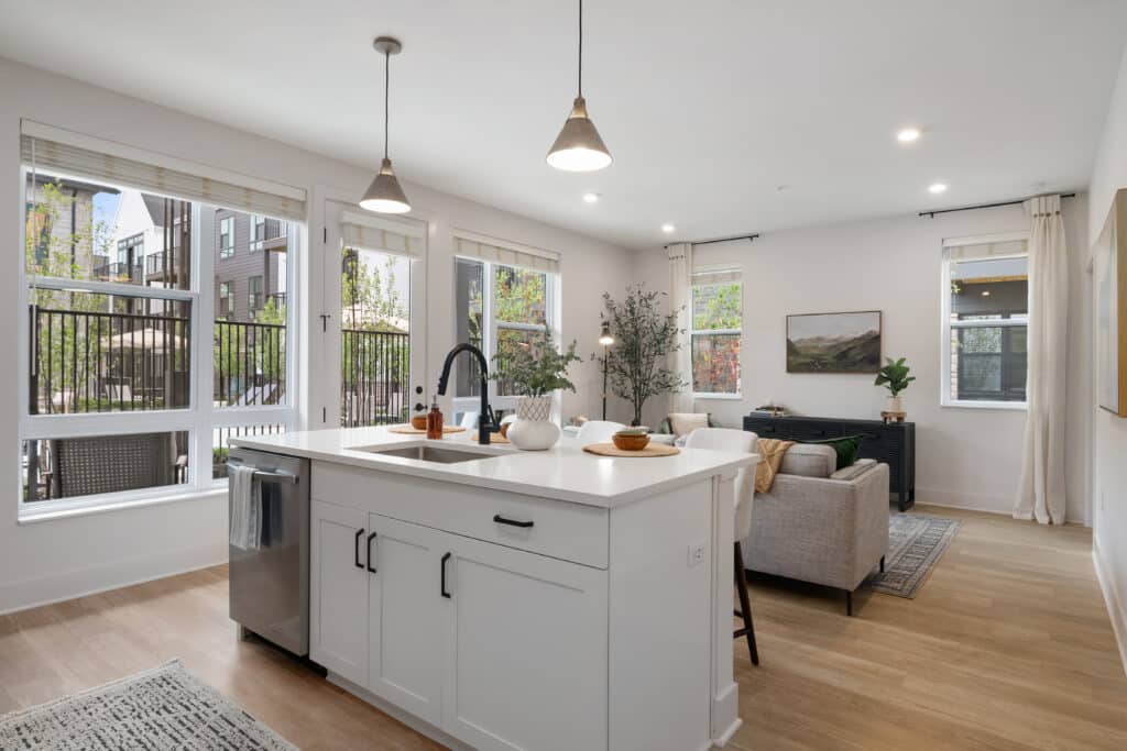 Modern open-concept one bedroom apartment featuring a kitchen and living room with large windows, a central island, neutral furniture, and abundant natural light.