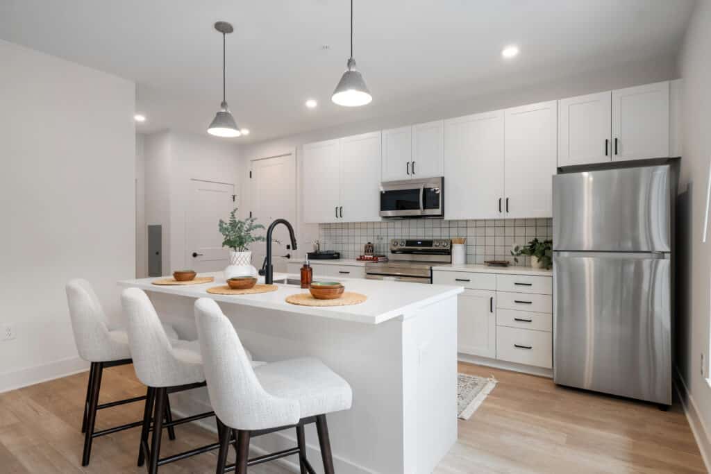 Modern kitchen in a stylish One Bedroom Apartment features white cabinets, stainless steel appliances, a spacious island with three upholstered bar stools, and elegant pendant lighting overhead.