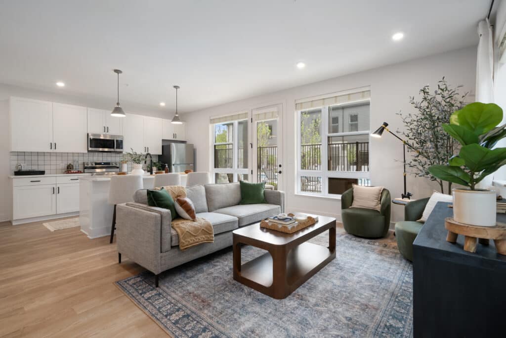 Modern open-plan living room and kitchen in a one bedroom apartment with neutral decor, large windows, gray sofa, green chairs, and wooden accents on light wood flooring.