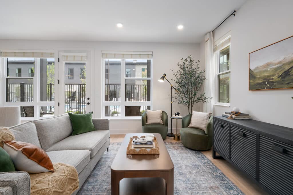 Modern living room in a one bedroom apartment with a gray sofa, wooden coffee table, two green chairs, large windows, and a sideboard with decor, featuring neutral tones and natural light.
