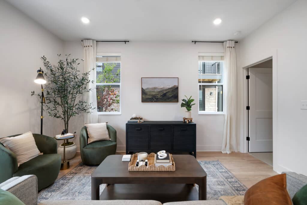 A modern living room in a one bedroom apartment with green chairs, a gray sofa, a wooden coffee table, and a black sideboard. Natural light from two windows with white curtains brightens the space, complemented by wall art and a potted plant.