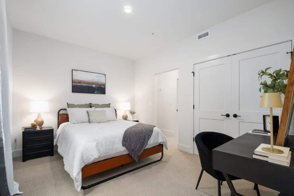 A neatly arranged one bedroom apartment featuring a bed, two nightstands with lamps, a desk with a chair, and white walls with minimal decor.