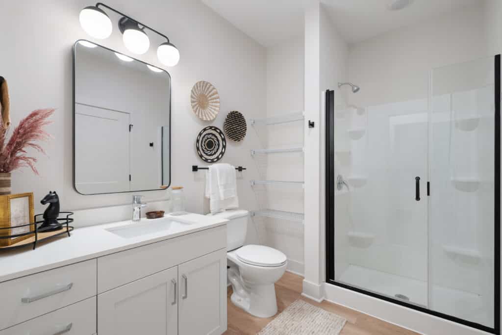 Modern bathroom in a One Bedroom Apartment featuring a white vanity, rectangular mirror, toilet, glass shower, wall-mounted lights, wicker wall art, and shelves. Light wood floor and neutral decor accents complete the space.