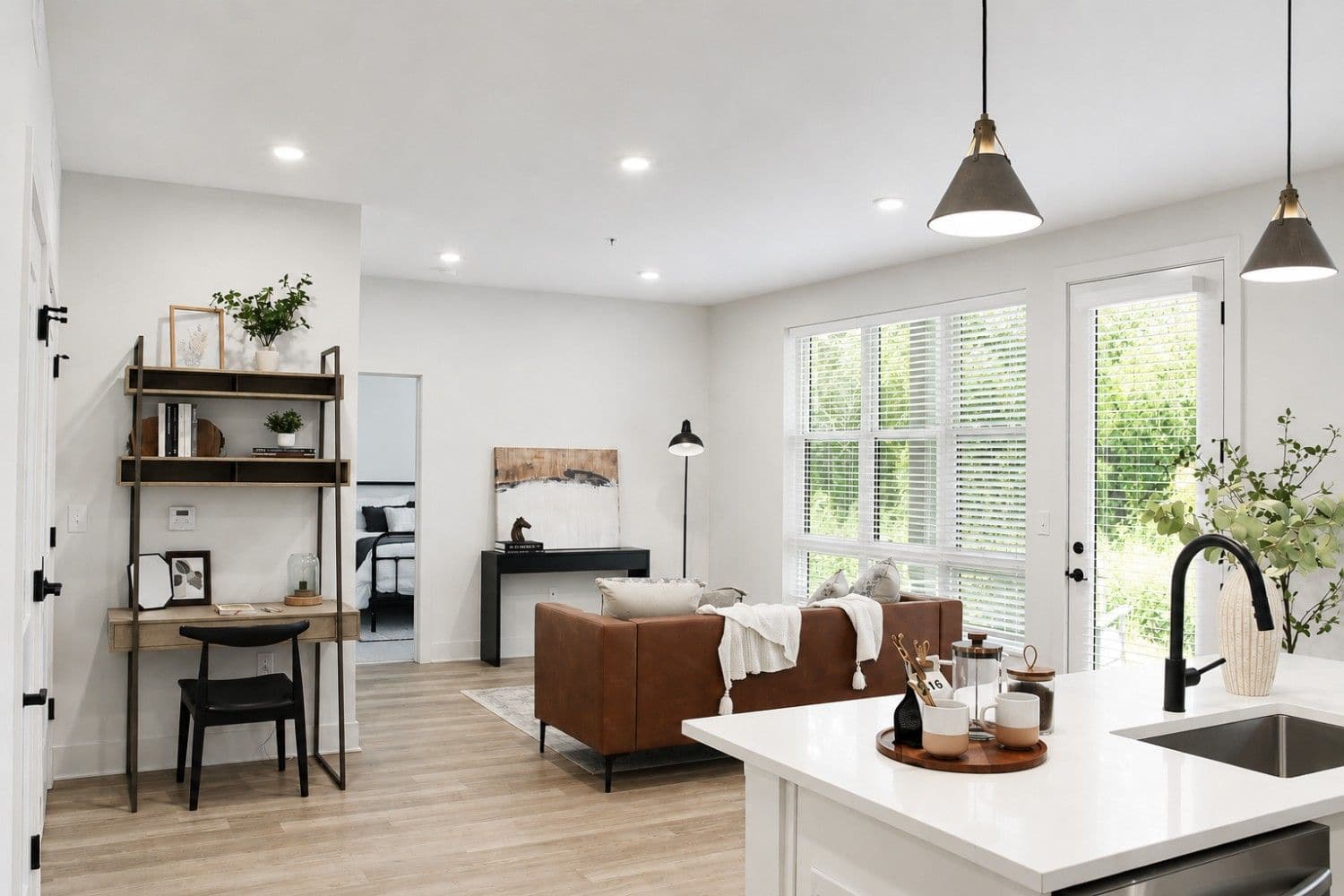 Open-plan living area with a brown sofa, desk, and shelves; white walls, large windows, and a kitchen island with modern lighting fixtures.