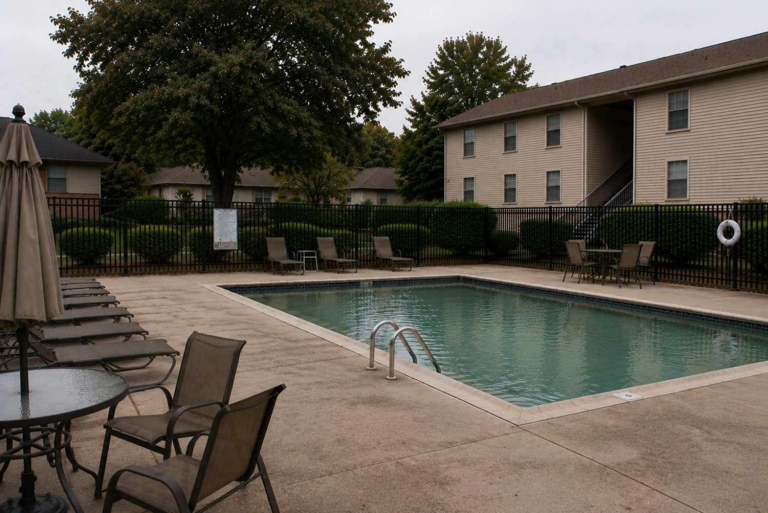 Outdoor swimming pool at an apartment complex, surrounded by lounge chairs, tables, and a black metal fence; trees and beige two-story buildings in the background.