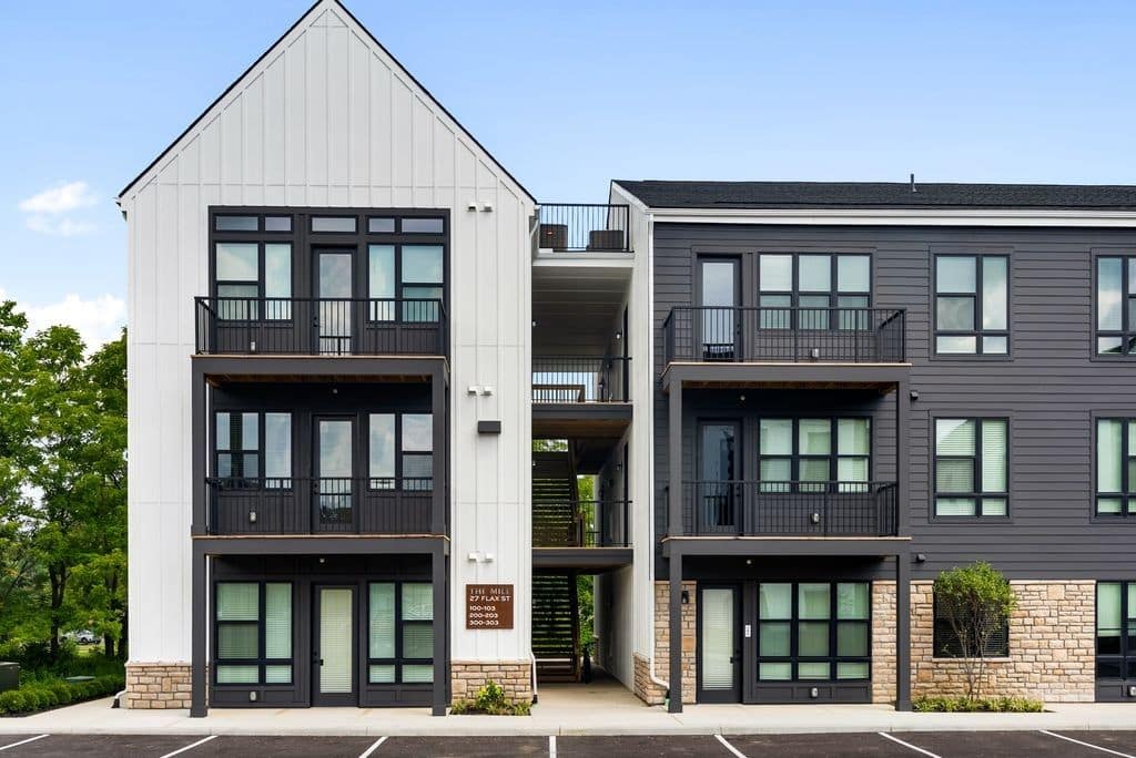 Modern three-story apartment building with contrasting white and gray siding, large windows, balconies, and a central open stairwell, set against a clear sky with empty parking spaces in front.