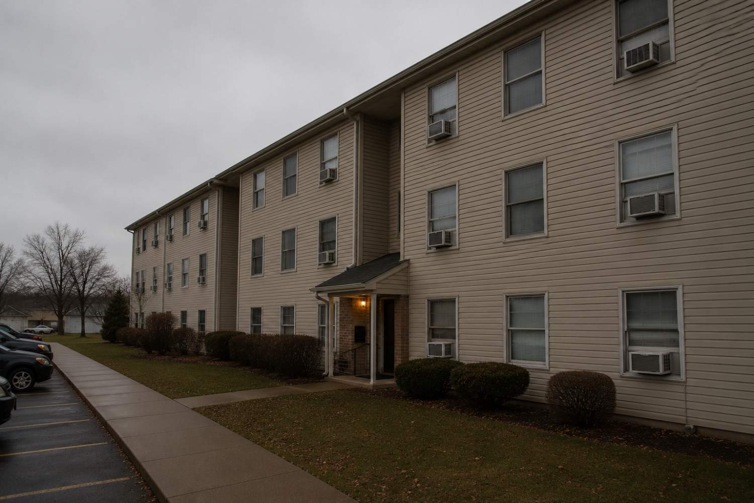 A three-story beige apartment building with multiple windows and window air conditioners, seen on an overcast day. Bushes line the front and several cars are parked to the left.