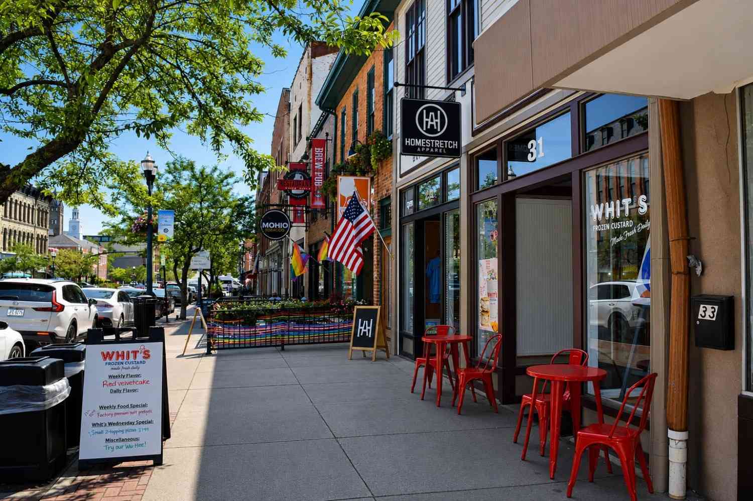 A city sidewalk with outdoor seating, storefronts, and American flags. Red chairs and tables are in front of Whit's, and signage for several businesses is visible. Trees provide some shade.