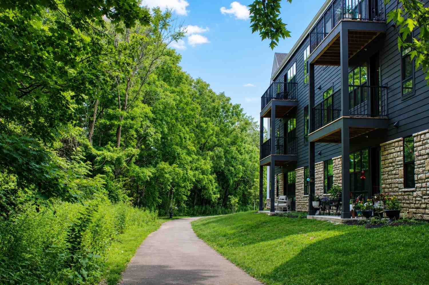 A paved path runs alongside a modern apartment building with balconies and patio furniture, bordered by lush green trees and grass under a blue sky.