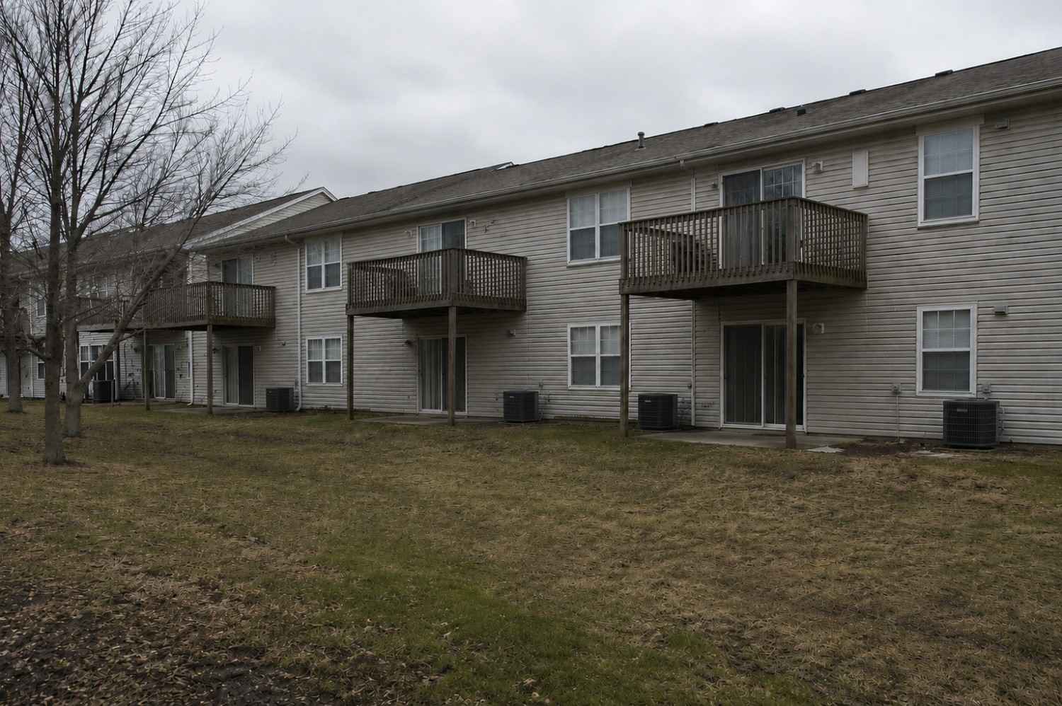Two-story apartment building with wooden balconies, sliding glass doors, and air conditioning units, on a cloudy day with bare trees and a patchy lawn.