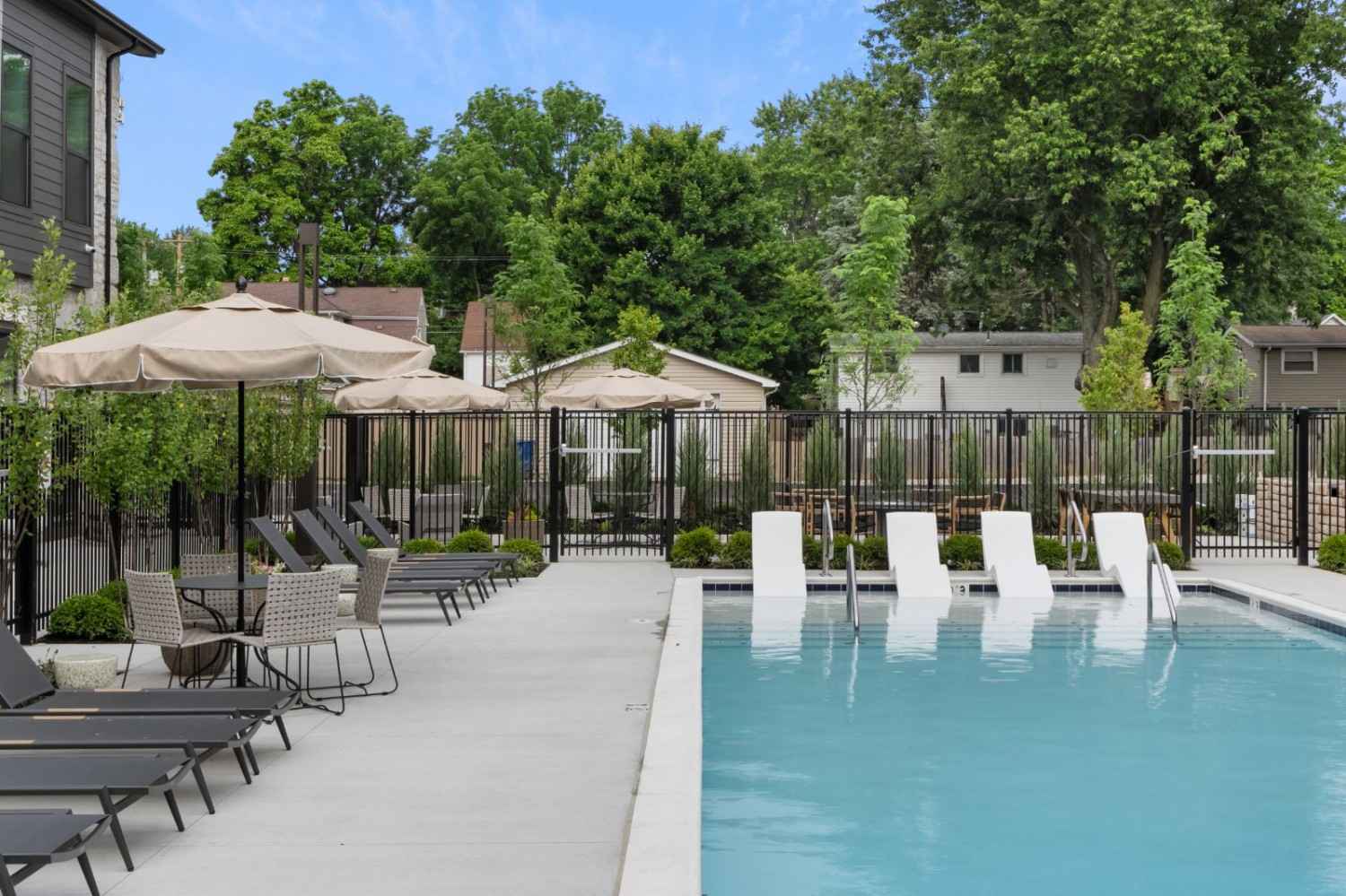 Outdoor pool with white lounge chairs partially submerged in water, black metal fence, shaded seating area with umbrellas, and leafy trees in the background.