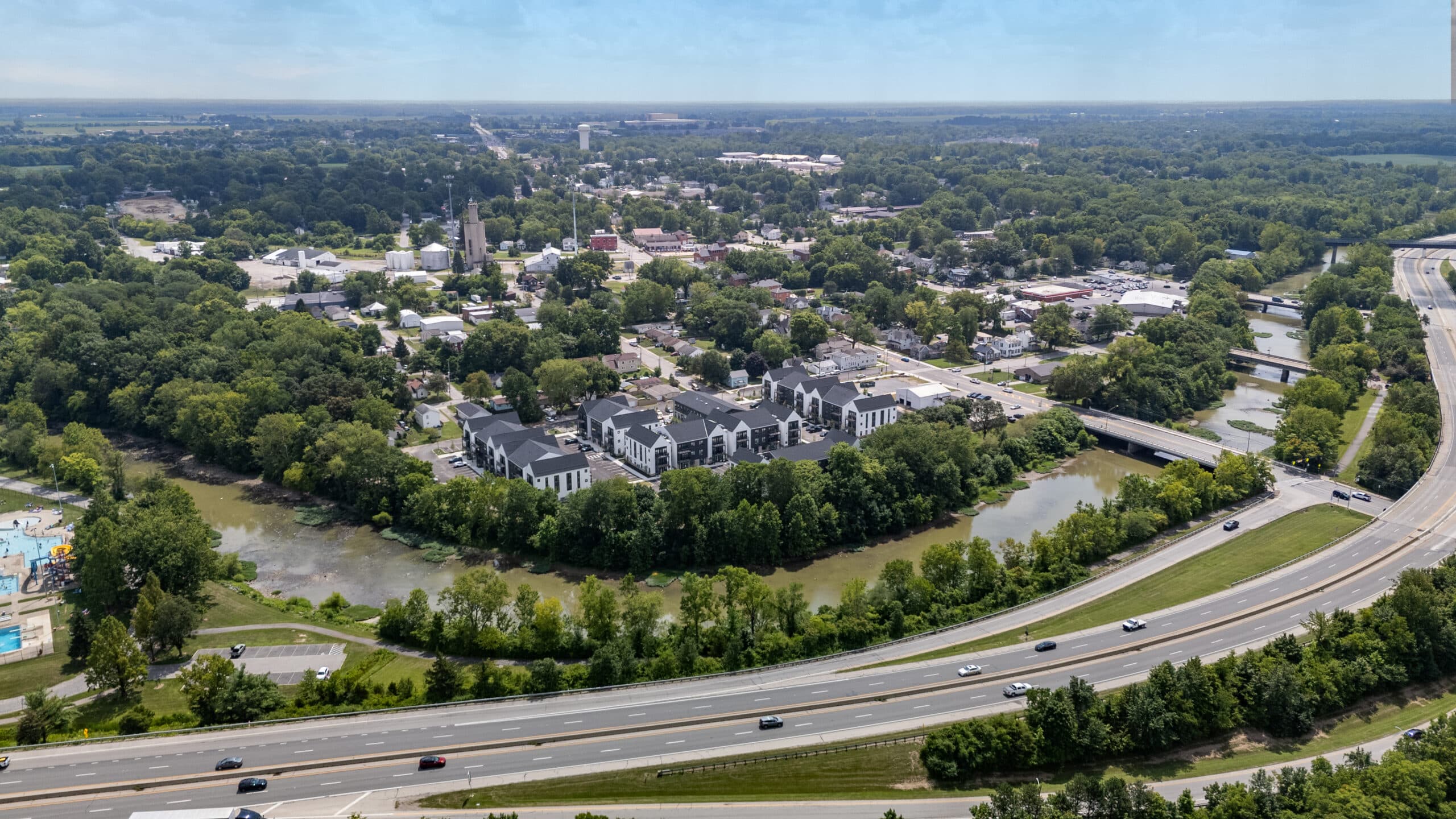 Aerial view of a suburban area with Delaware luxury apartments, a river, dense trees, and cars on nearby highways under a clear sky.